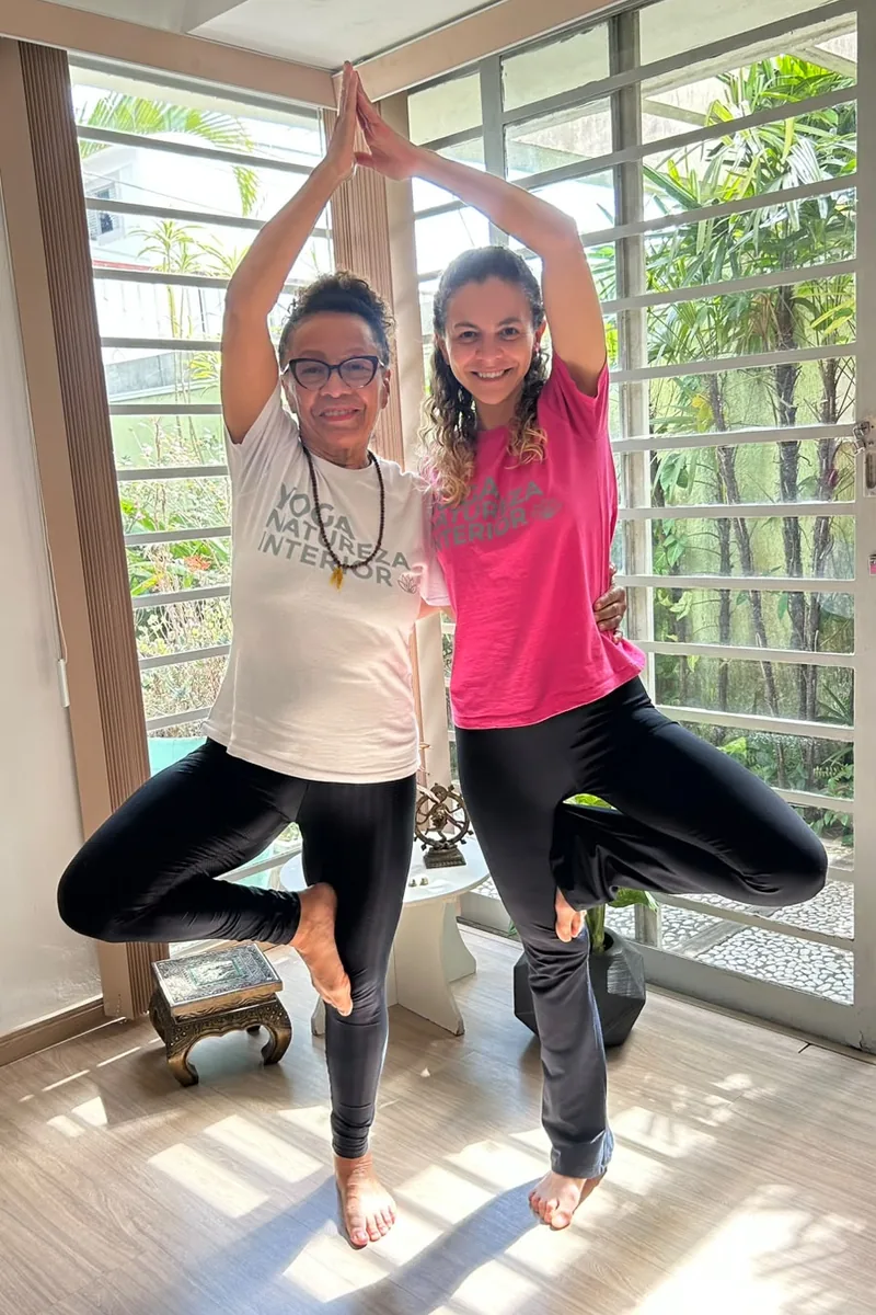 Rô e Káccia praticando a postura da árvore (Vṛkṣāsana) em uma sala de Yoga do Natureza Interior